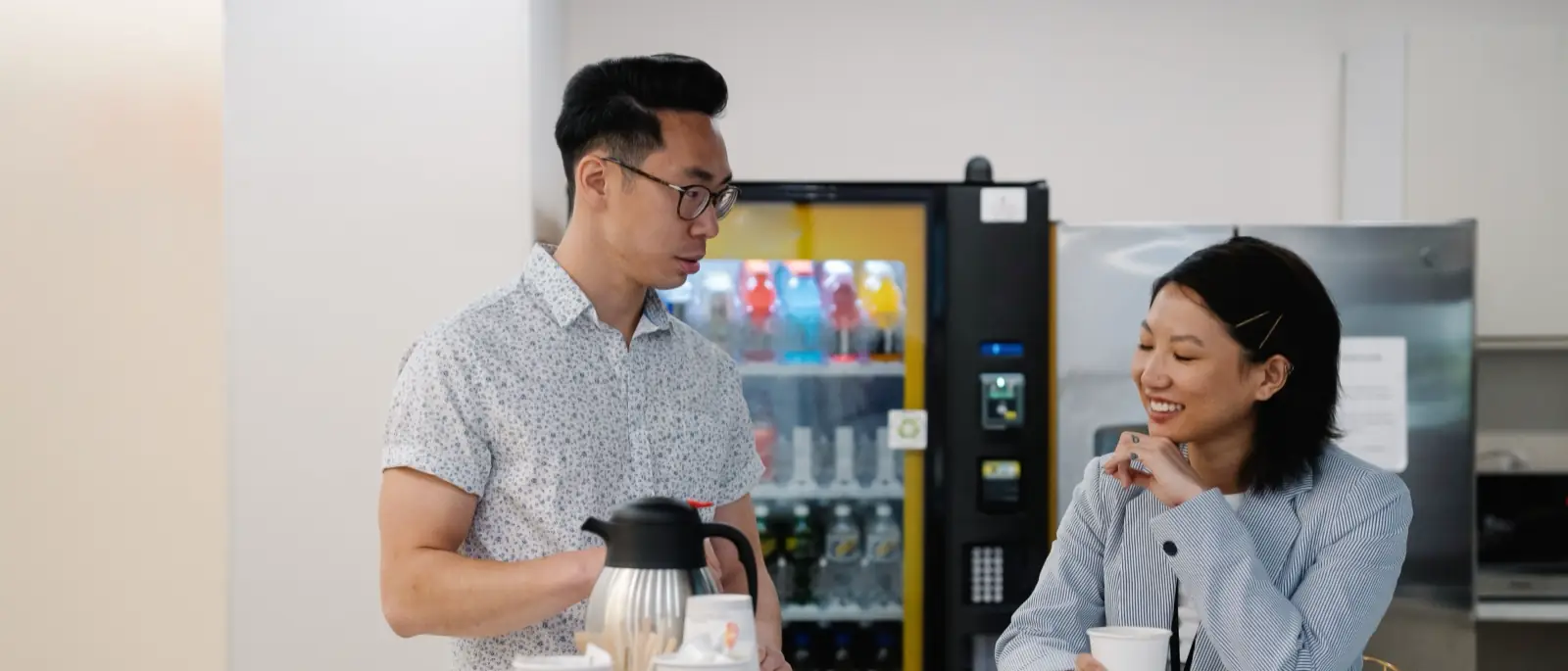 Dos compañeros de trabajo conversan junto a la barra del área de descanso mientras uno sirve café, con la máquina expendedora encendida al fondo
